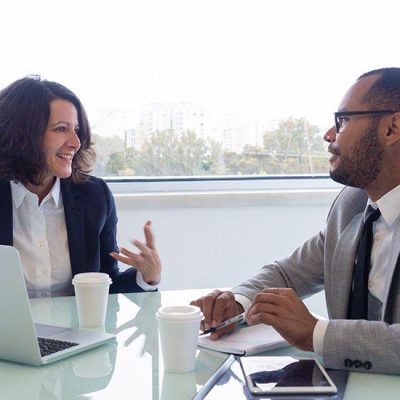 Happy female mentor explaining work issues to male intern. Business man and woman sitting at meeting table with open laptop and talking. Partnership concept